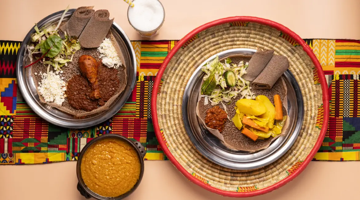 Zwei Injera-Teller zum Teilen auf bunten Läufern: Doro Wat, Gemüse, Salat und frischer Käse, dazu ein geflochtener Mesob