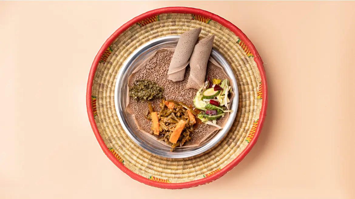 Veganes Injera-Tablett zum Teilen: Gomen, Atkilt-Gemüse und Salat auf frischer Injera im geflochtenen Korb